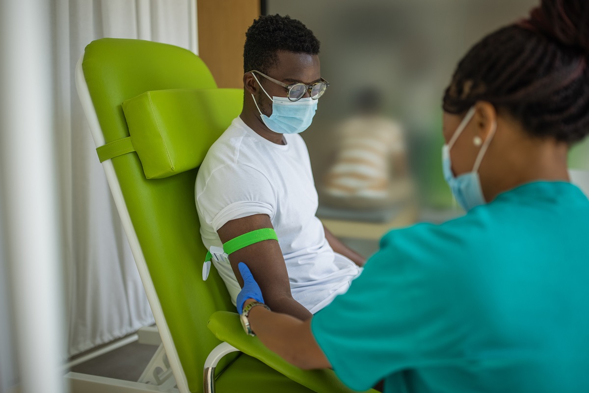 A man sits in a chair at a medical clinic, with a tourniquet on his right arm. A nurse, facing the patient, holds the arm.