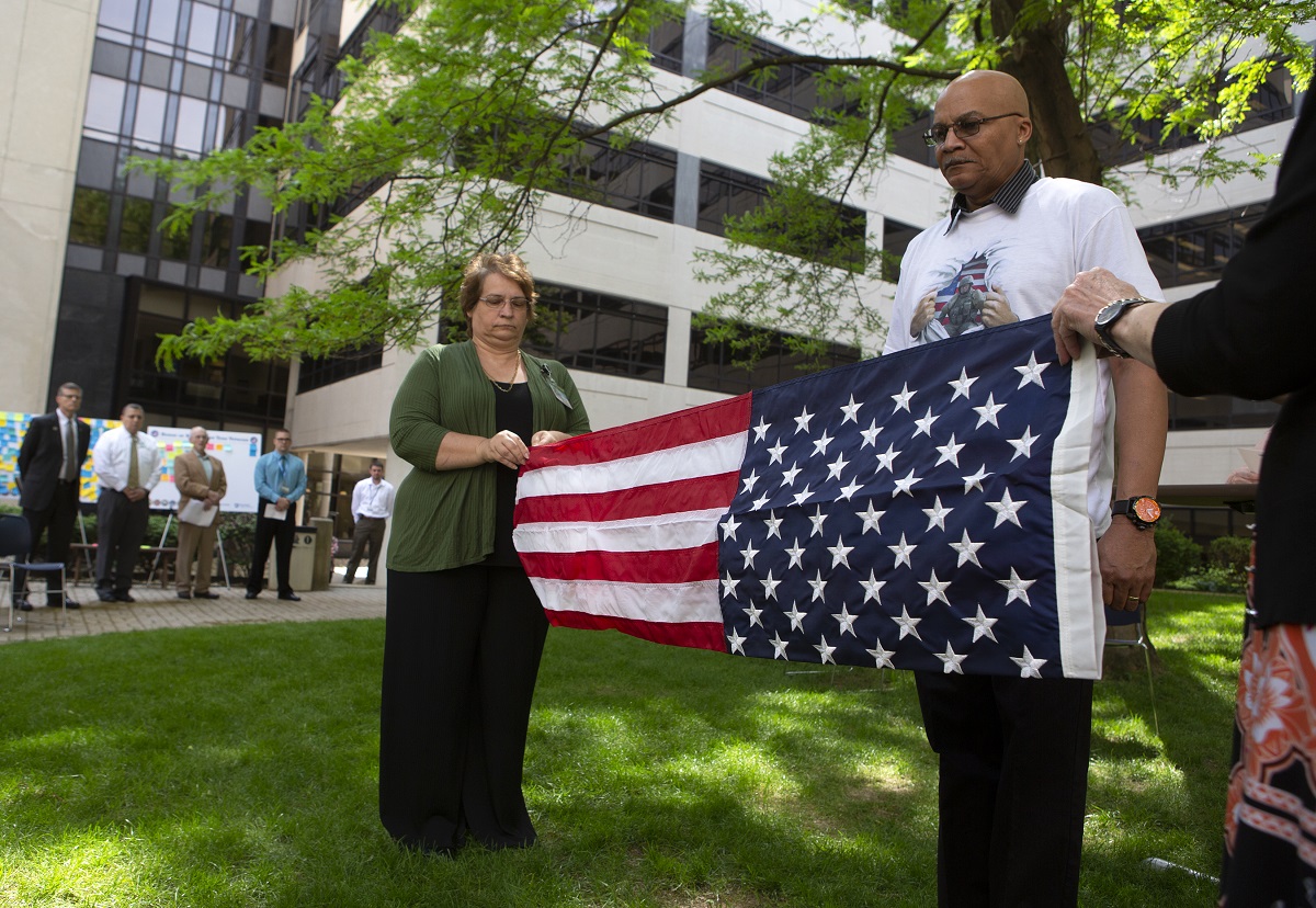 In a courtyard, two people fold an American flag. A man stands at attention in front of the flag. Behind them, a row of people look on from a walkway.