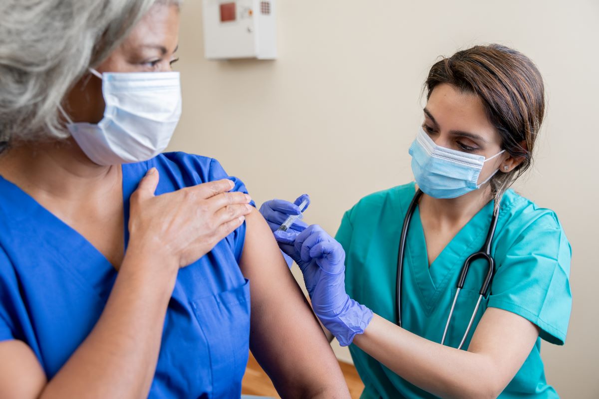 Health care professional in scrubs and medical mask administering a shot to another health care professional in scrubs and a medical mask.