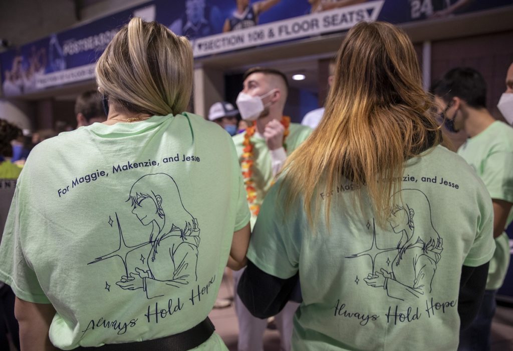 Two women shown from behind in the foreground wear T-shirts with the words For Maggie, Makenzie, and Jesse. Written on the them. The shirt includes a design of a woman holding stars. The text beneath that says Always Hold Hope. Jesse king, wearing flowers, can be seen in the background.