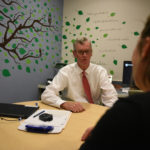 Dr. George Blackall, PsyD, MBA, is seen sitting at table counseling a female patient. He is looking directly at her. The patient Is facing Dr. Blackall and her face is not visible. The room has a decal of a tree on its walls.