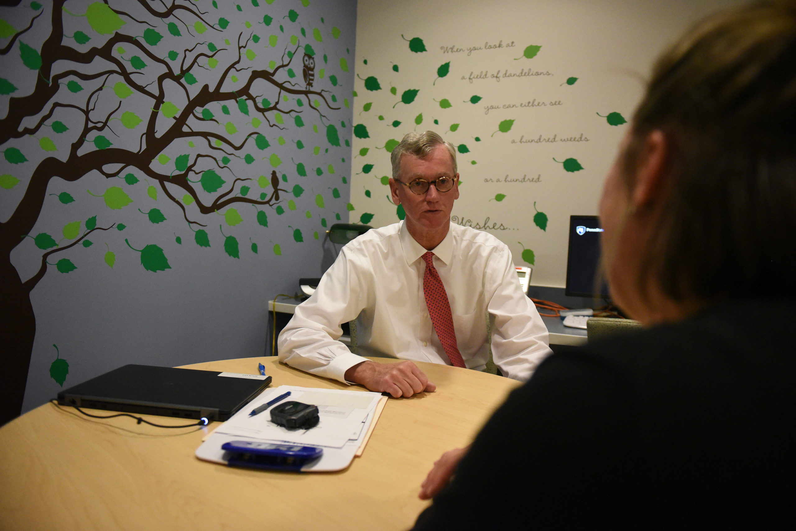 Dr. George Blackall, PsyD, MBA, is seen sitting at table counseling a female patient. He is looking directly at her. The patient Is facing Dr. Blackall and her face is not visible. The room has a decal of a tree on its walls.