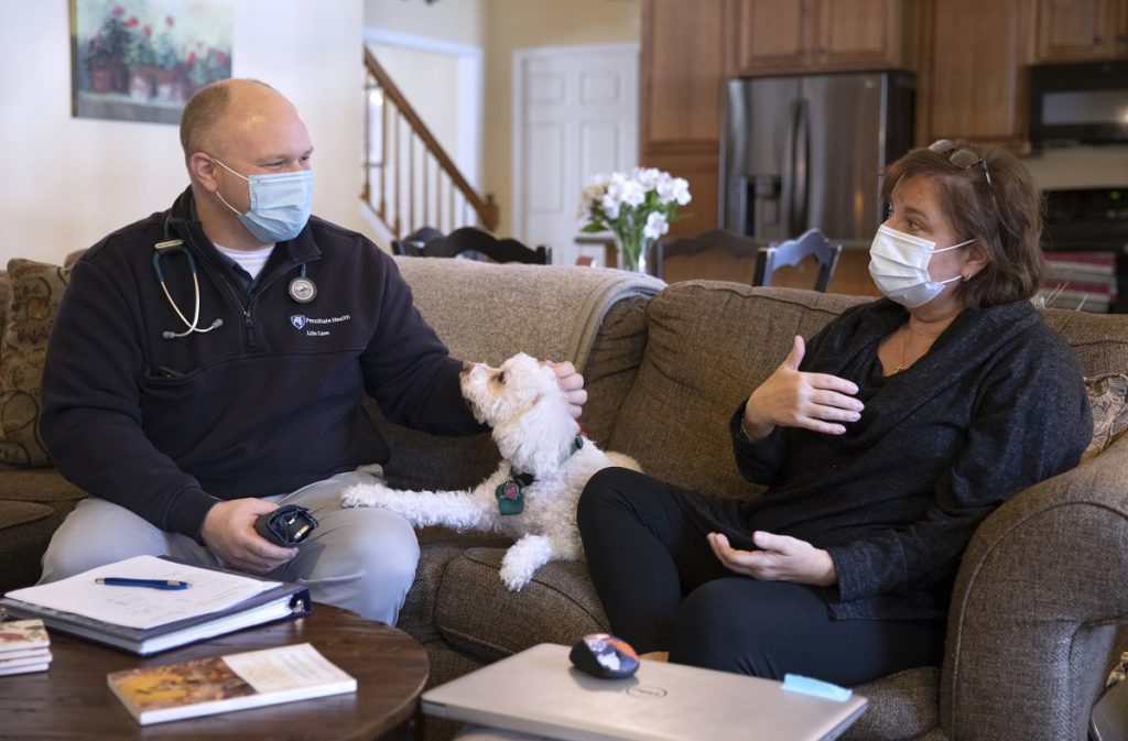 Kate Warnigiris, who has short hair with glasses on the top of her head and wears a shirt, pants and face mask, gestures as she talks with Loren Miller on a couch in her living room. Miller, dressed in a Penn State Health jacket with a stethoscope around his neck and a face mask, pets a dog on the couch as he listens.