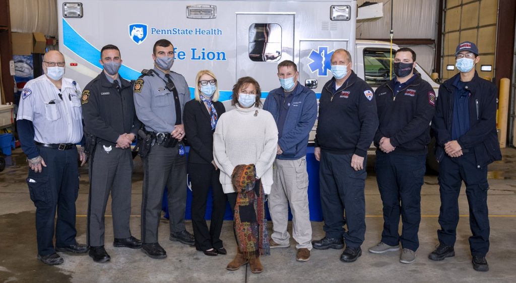 Kate Warnigiris, wearing a bulky sweater, jeans and a mask, is surrounded by eight people who helped save her life during a cardiac arrest. Crew members, who are wearing uniforms from their respective agencies and masks, stand in front of a Life Lion ambulance in a garage.