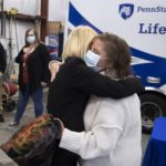 Kate Warnagiris, who has short hair and wears a bulky sweater and mask, embraces Dr. Monica Corsetti, shown from the back, who is wearing a jacket and has short hair. A Life Lion ambulance and crew members are standing are in the background.