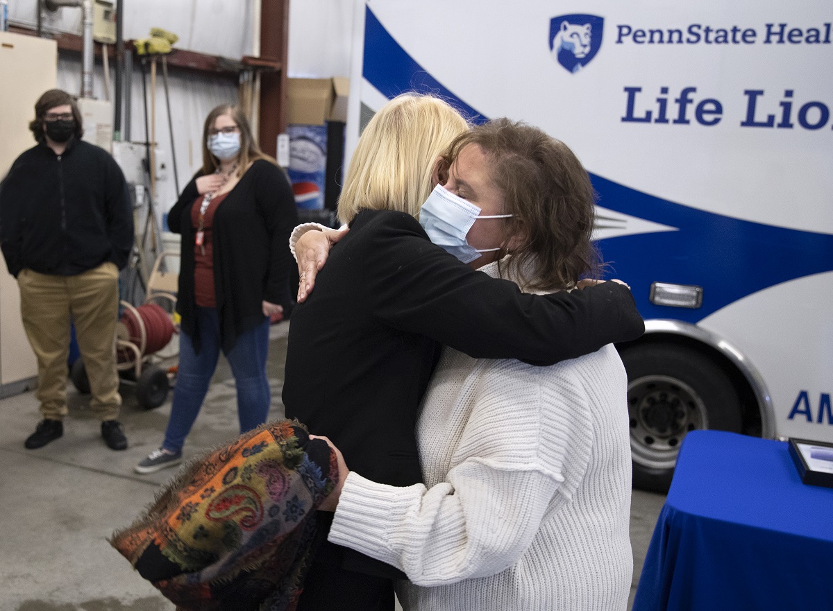 Kate Warnagiris, who has short hair and wears a bulky sweater and mask, embraces Dr. Monica Corsetti, shown from the back, who is wearing a jacket and has short hair. A Life Lion ambulance and crew members are standing are in the background.