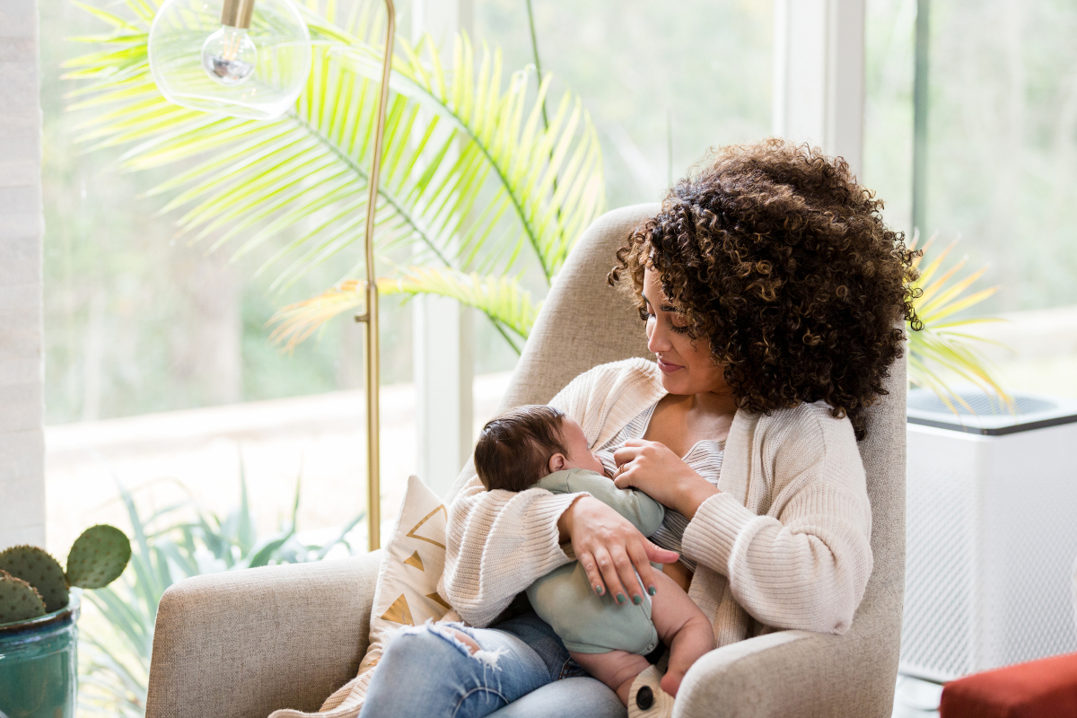 A woman sits in a chair, breastfeeding her baby. A lamp and some plants are in the background, as is a large window.