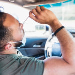 A man sitting in the driver’s seat of a car uses his right hand to insert a nasal swab into his nose, with his head tilted backward slightly.
