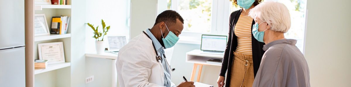 In a doctor’s office, a doctor is seated and writing in a chart while sitting across from a seated, elderly female patient. A younger woman stands by her side. All are wearing face masks.