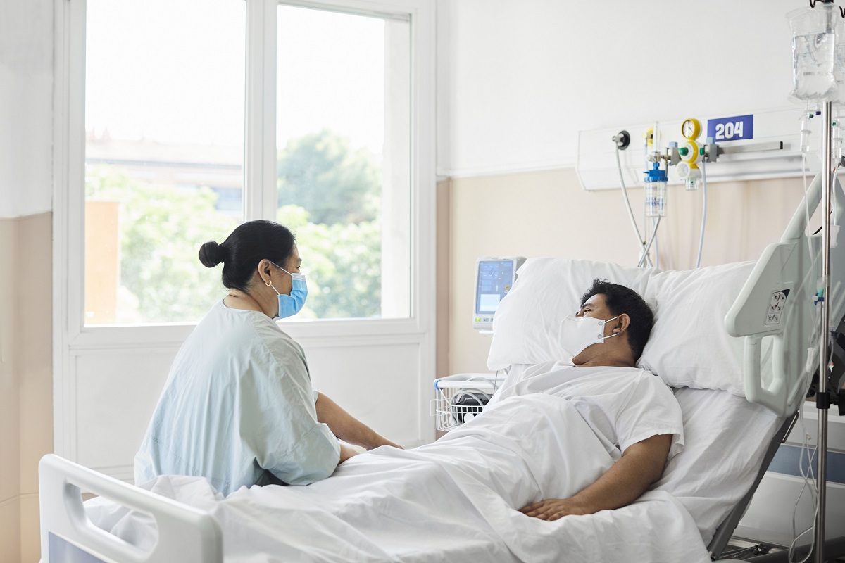 A woman sits at the edge of a hospital bed. A man lies in the bed. Both wear masks.