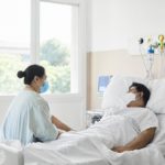A woman sits at the edge of a hospital bed. A man lies in the bed. Both wear masks.