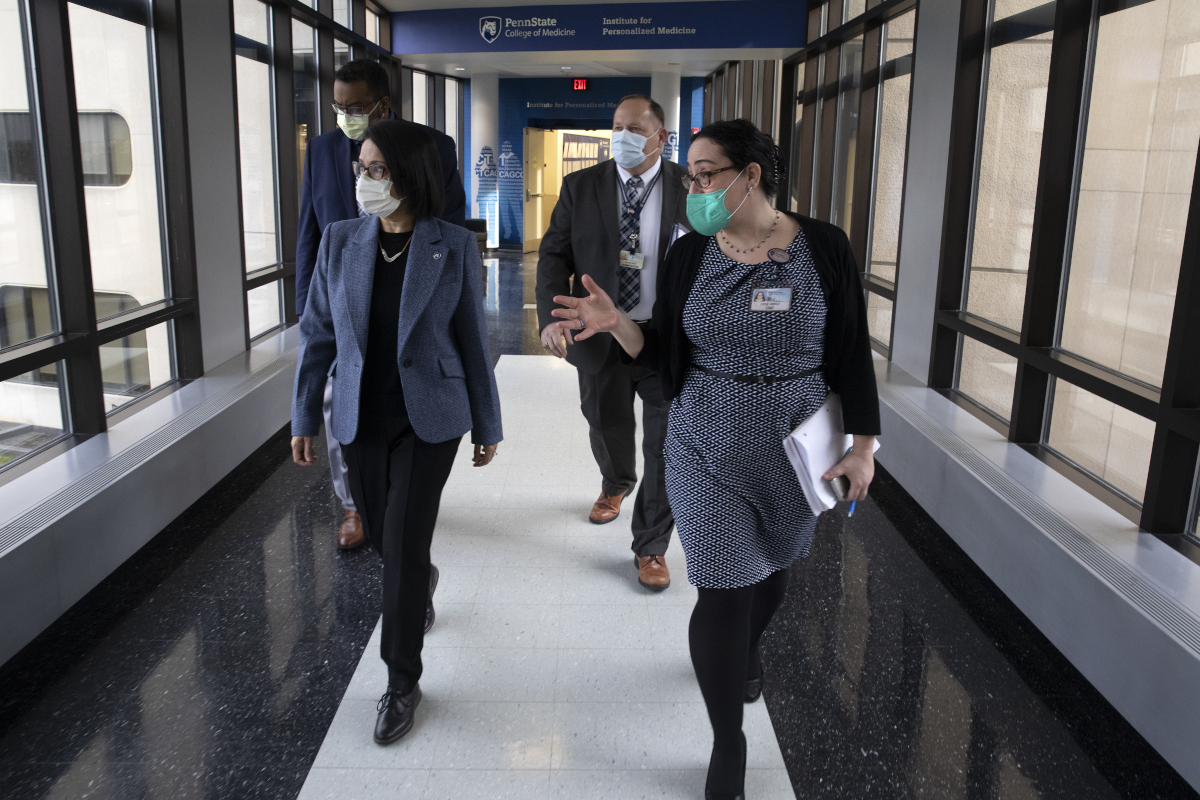 Four people – two women followed by two men – walk down a hallway with signage that reads “Penn State College of Medicine” and “Institute for Personalized Medicine.” As they walk, they look to their right, out of a set of windows.