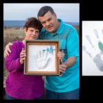 Wendy Gilbert, who has short hair and wears a bright top and jean, and Ronald Gilbert, who wears a polo shirt and jeans, hold a framed shadow box containing a resin handprint and 3D heartbeat. They are standing outside with a field in the background. Dylan’s handprints are beside them.