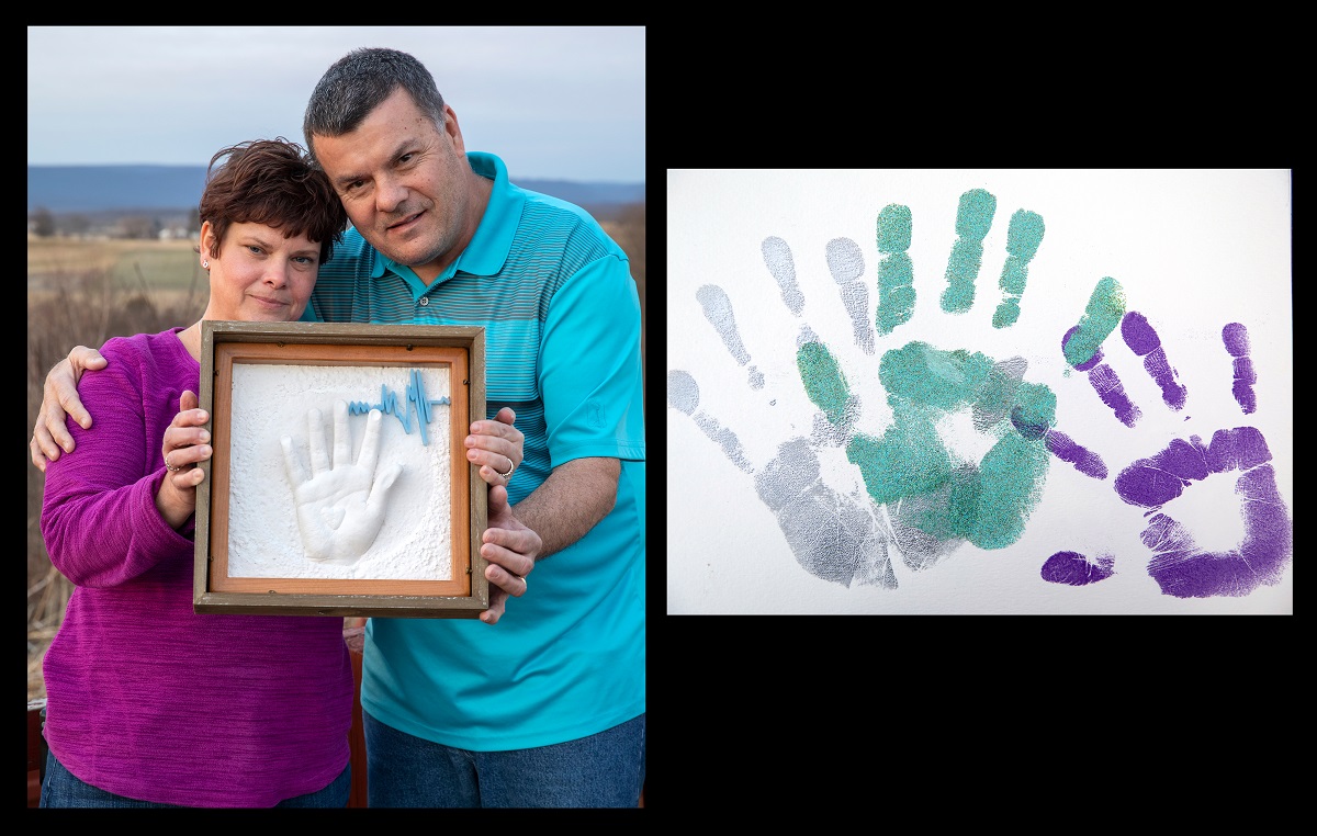 Wendy Gilbert, who has short hair and wears a bright top and jean, and Ronald Gilbert, who wears a polo shirt and jeans, hold a framed shadow box containing a resin handprint and 3D heartbeat. They are standing outside with a field in the background. Dylan’s handprints are beside them.