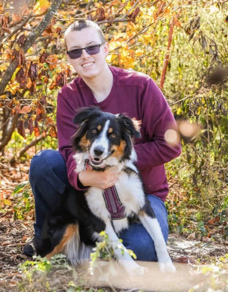 Dylan Gilbert smiles and kneels in front of a tree with his Australian shepherd, Mocha, who is brown, white and black. Dylan wears glasses, a shirt and jeans. 