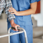 An elderly man leans on a walker with the assistance of a health care worker in scrubs.