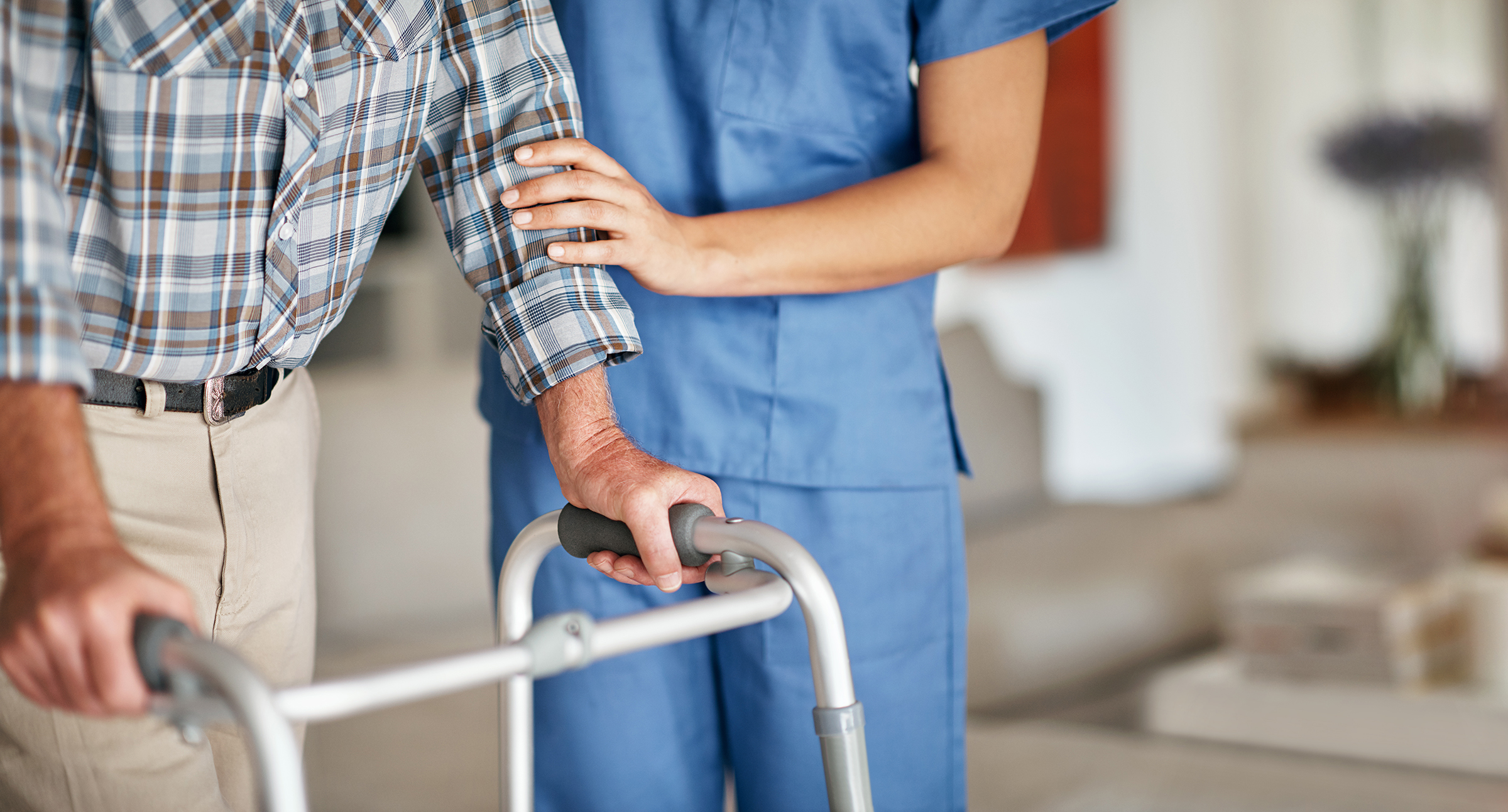 An elderly man leans on a walker with the assistance of a health care worker in scrubs.