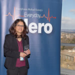 Dr. Gayatra Mainali, wearing professional attire, stands in front of a large pull-up banner with the words “Pennsylvania Medical Society’s Everyday Hero.” Buildings, trees and mountains are in the background, outside a window.