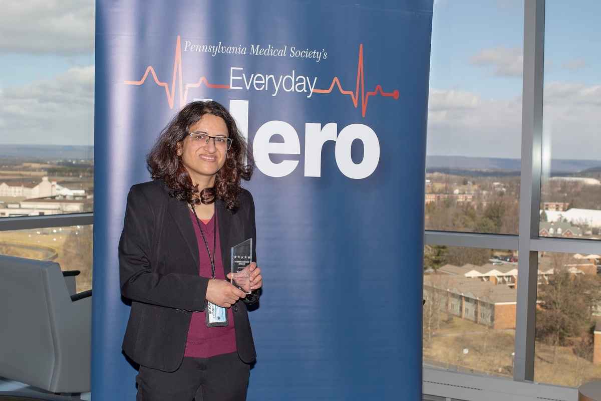 Dr. Gayatra Mainali, wearing professional attire, stands in front of a large pull-up banner with the words “Pennsylvania Medical Society’s Everyday Hero.” Buildings, trees and mountains are in the background, outside a window.