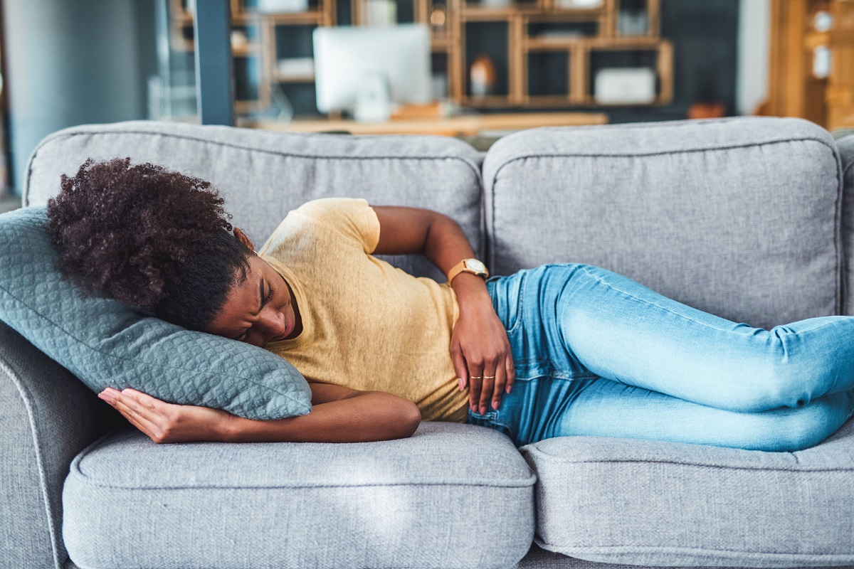 A young woman experiencing abdominal pain lies on the sofa at home.