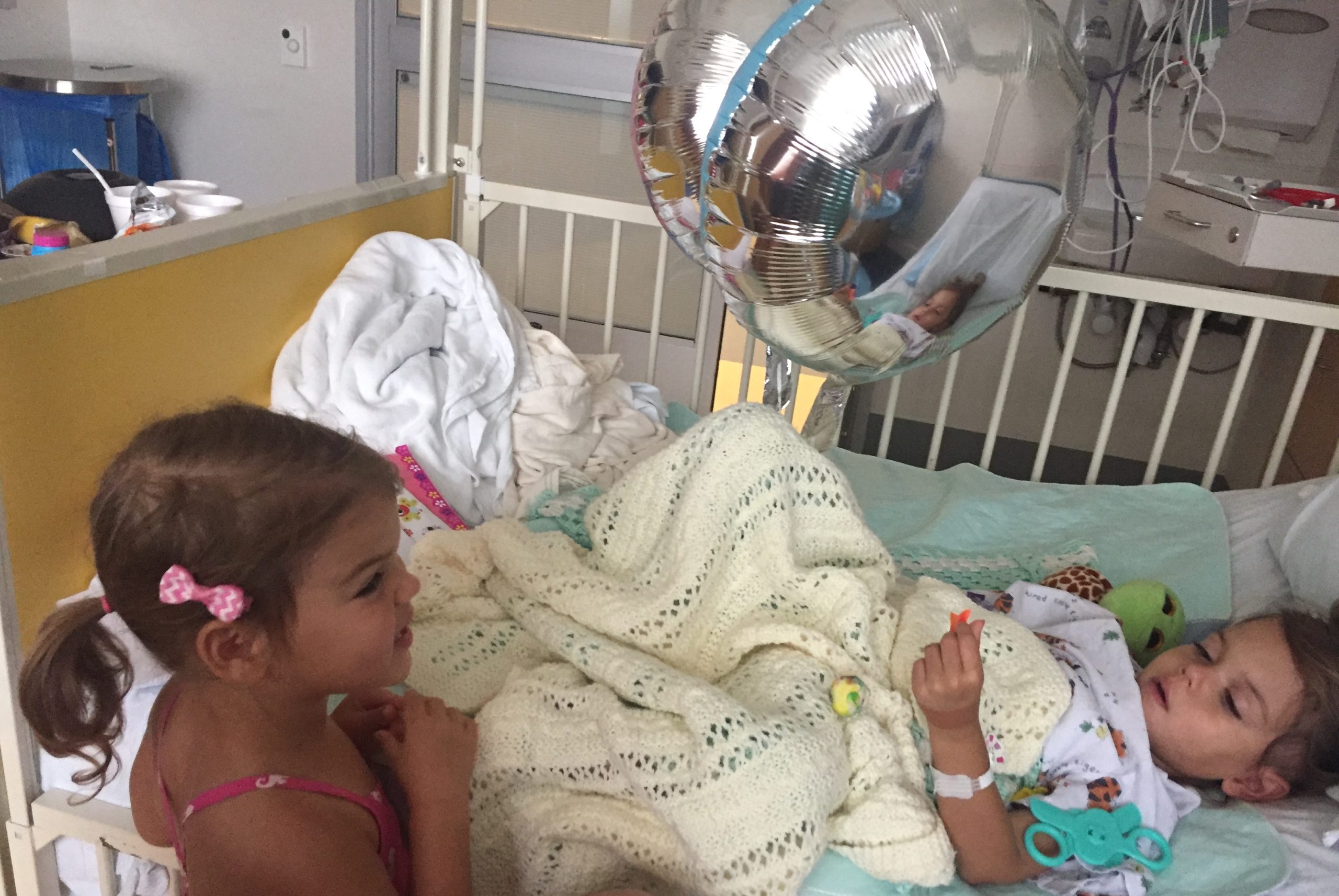 A young girl with pigtails and a ribbon in her hair visits another young girl in a hospital bed.