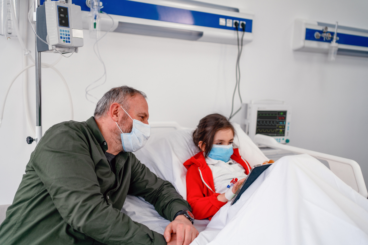 A young girl sits up in her hospital bed, playing on a tablet computer. She’s hooked up to medical equipment. A man seated at the side of the bed looks at the screen with her.