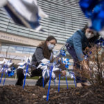 Two women kneel down and place plastic pinwheels into the ground by hand. Several pinwheels are in the foreground, close to the camera. A building with the words “Children’s Hospital” is in the near background.