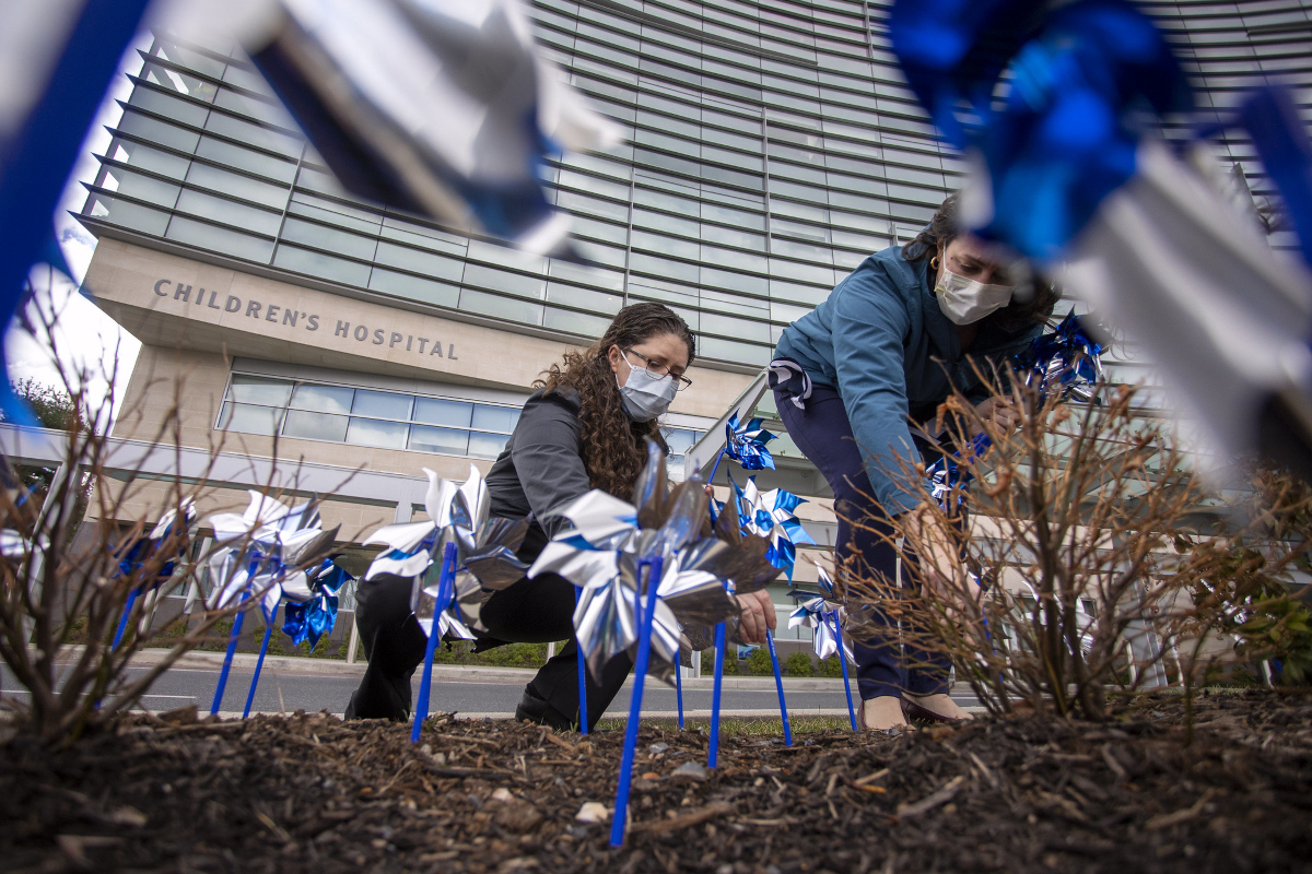 Two women kneel down and place plastic pinwheels into the ground by hand. Several pinwheels are in the foreground, close to the camera. A building with the words “Children’s Hospital” is in the near background.