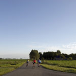 Image of bicyclists pedaling down a road in a rural area. Trees and farmland are in the distance.