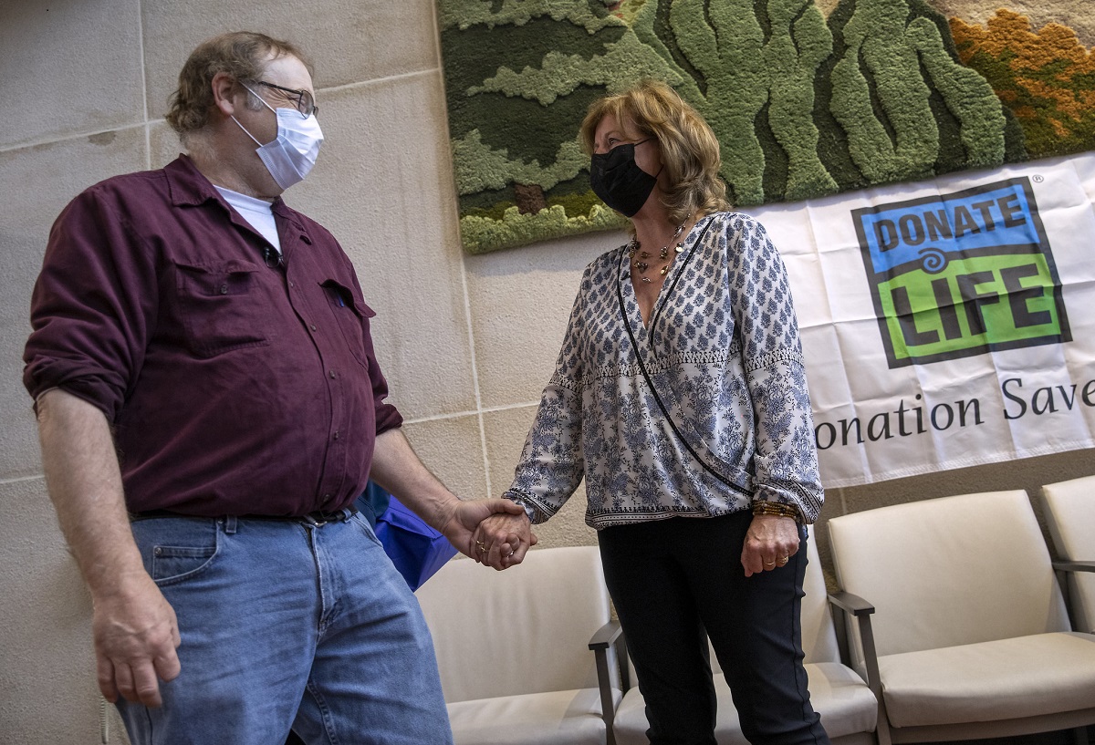 Dwayne Weller, who wears glasses and a face mask and is dressed in a chamois shirt and jeans, clasps hands with Maureen Stathes, who has shoulder length hair and wears a face mask, blouse and pants. A “Donate Life” banner is on the wall behind them.