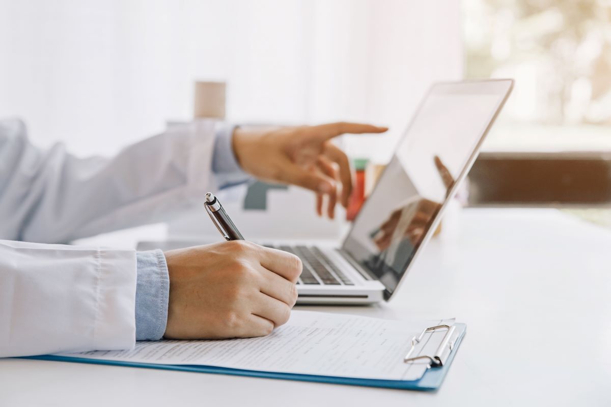 A healthcare professional wearing a white coat points at a laptop screen while writing on a paper on a clipboard.