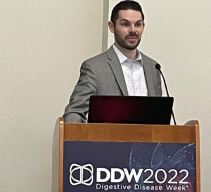 Dr. Mark Bundschuh, gastroenterology fellow, stands behind a podium with a microphone as he presents during the Digestive Disease Week conference.