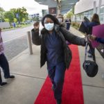 Nancy Ochieng, a registered nurse at Hershey Medical Center, dances her way to the main entrance on a red carpet during the kickoff of Nurses Week 2021. She is wearing a face mask, scrubs and a jacket and is carrying a bag. Three nurses holding up signs stand on each side of the carpet.