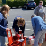 Four health care workers organize plastic containers full of syringes, which are placed at the edge of a parking lot. A car and bushes are in the background.