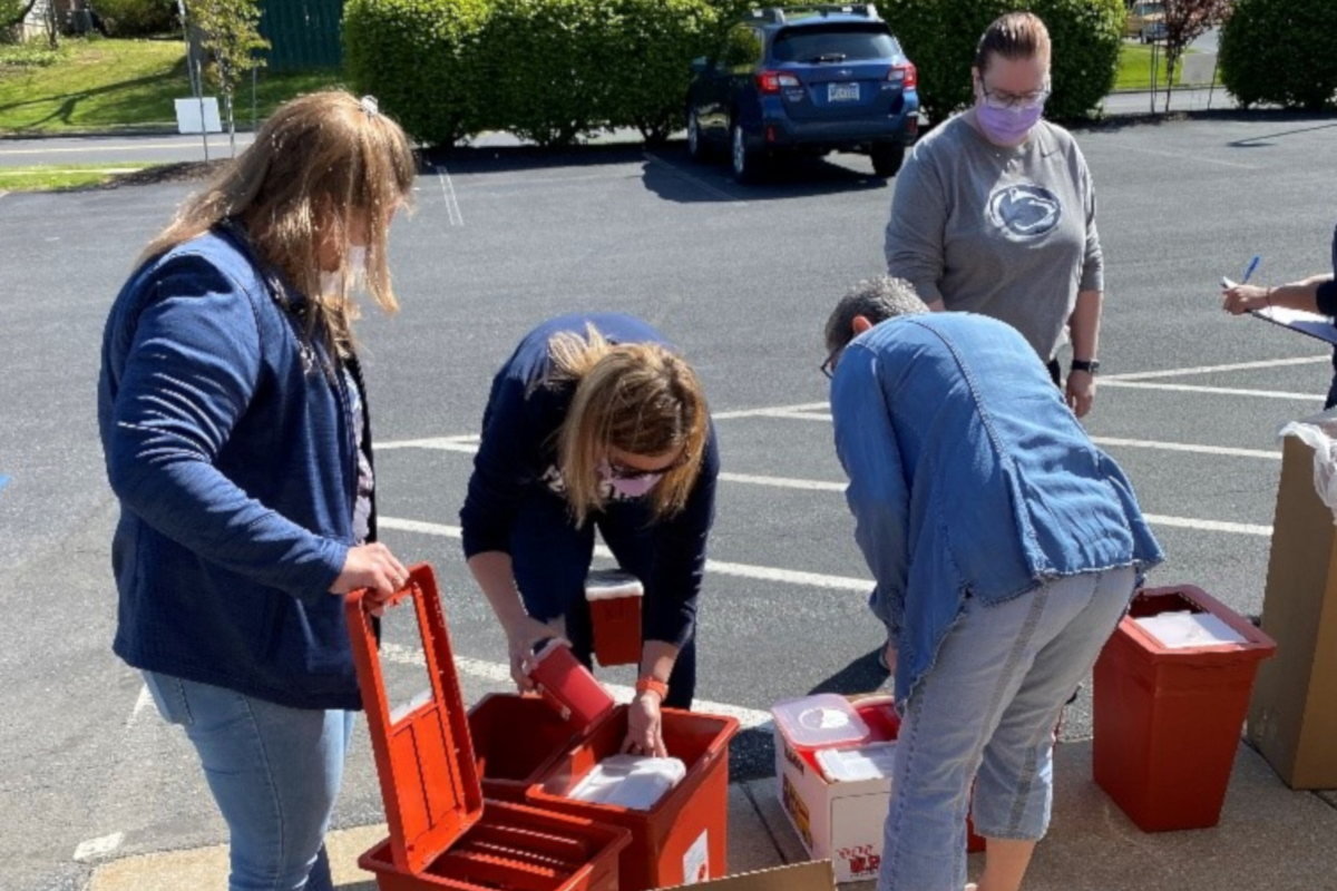 Four health care workers organize plastic containers full of syringes, which are placed at the edge of a parking lot. A car and bushes are in the background.