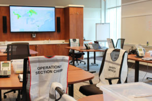 A conference room with tables and chairs placed throughout. Vests with job titles such as “operations sections chief” and “safety officer” are draped over many of the chairs. A window, easel, and large screen monitor are in the background.