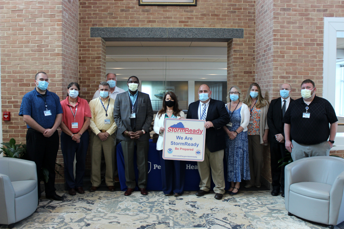 Eleven people stand in a row, posing with a certificate that reads “StormReady: We Are StormReady. Be Prepared.” A brick wall with an opening in the middle is in the immediate background.