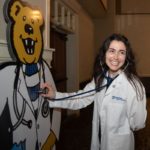 A woman wearing a physician assistant's coat holds up her stethoscope to a cardboard cutout of Penn State's Nittany Lion, also wearing a physician assistant's coat.