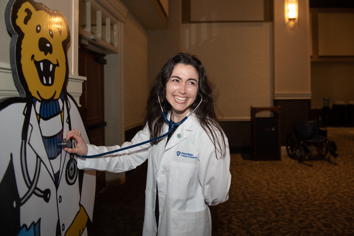 A woman wearing a physician assistant's coat holds up her stethoscope to a cardboard cutout of Penn State's Nittany Lion, also wearing a physician assistant's coat.