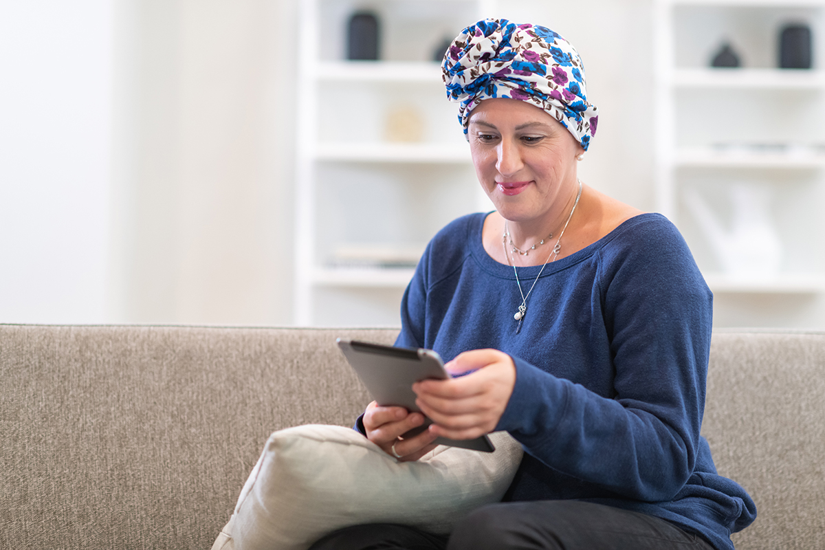 A female cancer patient uses a tablet while sitting on a couch.