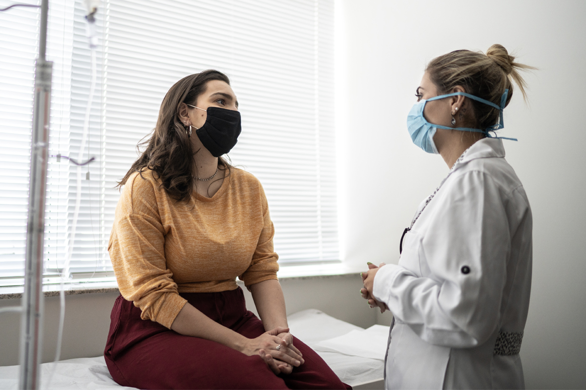 A woman speaks with her health care provider. She is seated, the provider is standing, a short distance away.