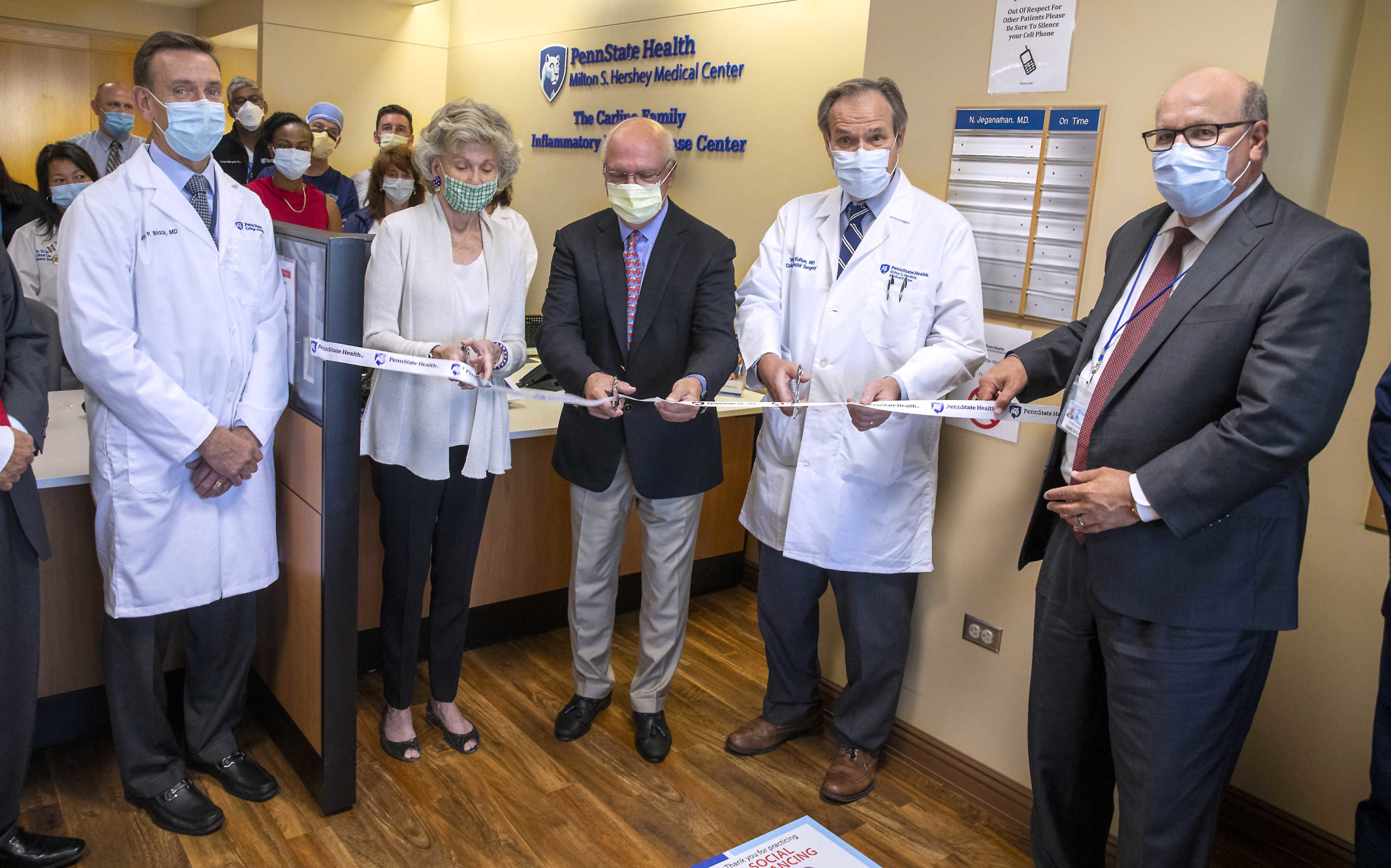 Five people in the foreground pose for a photo as they prepare to cut a ceremonial ribbon. A small group of people looks on in the background.