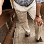 As he climbs stairs, an older man holds onto the bannister with his right hand and clutches his left knee with his left hand.