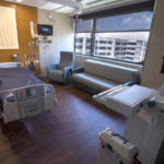 A hospital patient room with a bed, chair and sofa, as well as a headwall with various equipment and ports. A computer on a portable stand is in the foreground at right.