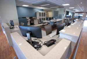Several computers and monitors line a long nurses’ station desk in a hospital unit. Additional desk space and other equipment is in the background.