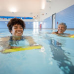 Two women exercise using kick boards in an indoor pool.