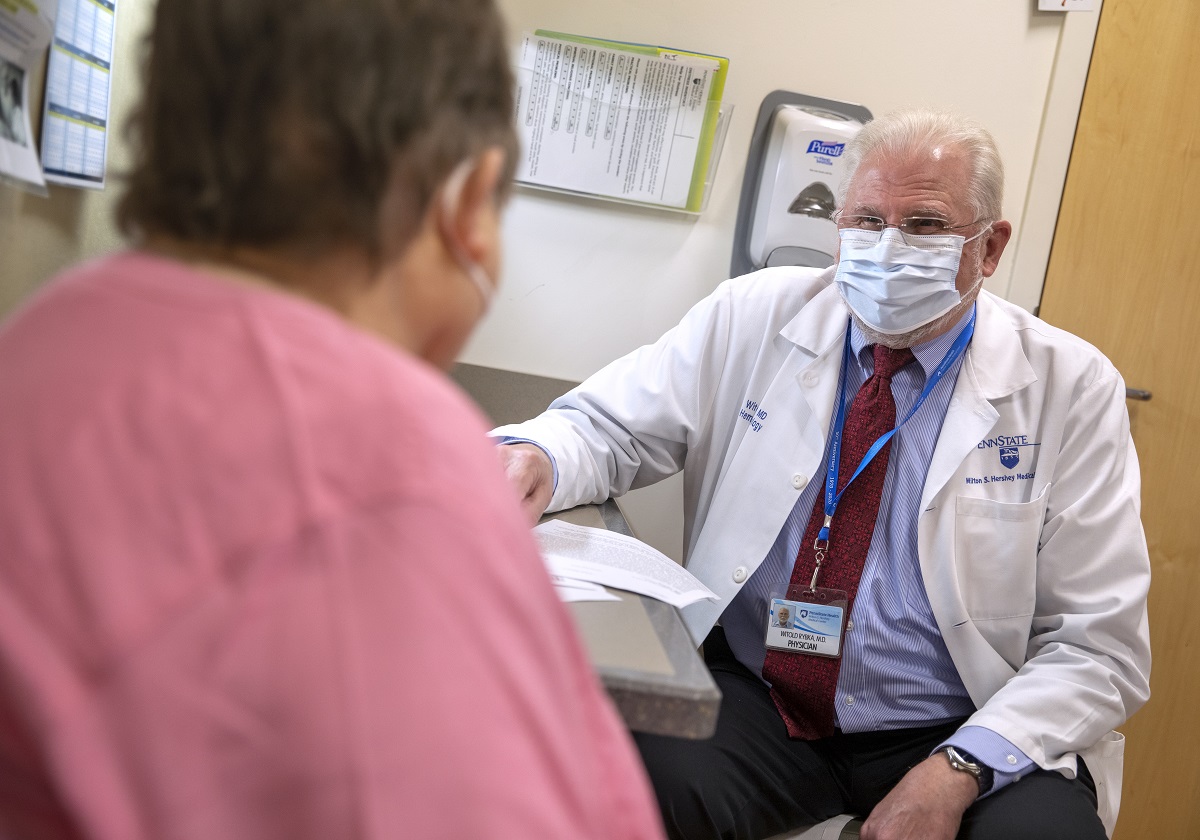 Dr. Witold Rybka, who wears a face mask, glasses and a white coat, sits in an exam room opposite a patient, who is seen from behind. A hand sanitizer dispenser and rack of papers are mounted on the wall behind him.