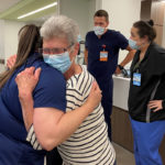 Former Hampden Medical Center patient Lora Lendvay hugs Hampden Medical Intensive Care Unit nurse Alexa Menapace. Lendvay is wearing a striped shirt, glasses and face mask. Menapace has long hair and is wearing scrubs. Behind them are a male nurse and female nurse, both wearing name badges.