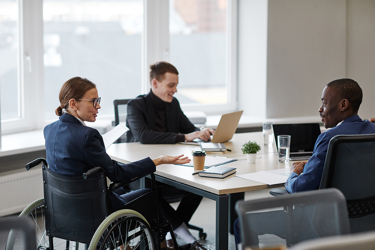 A woman in a wheelchair sits at a conference table with two men. She is wearing a suit jacket and glasses. The man to her left is smiling and typing on a laptop. The man to her right is smiling as he looks at her.