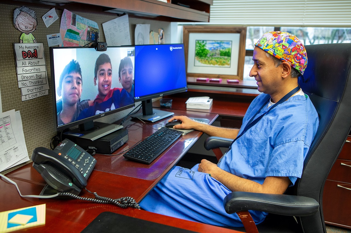 A doctor in scrubs smiles at images of three boys on his computer screen.